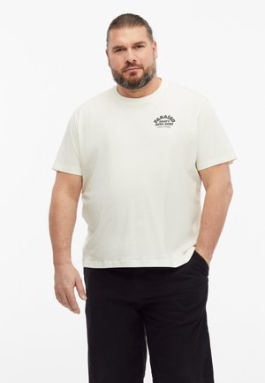 Mann mit Bart, der ein weißes T-Shirt mit kurzen Ärmeln und schwarzem Text auf der Brust sowie schwarze Hose trägt, eine Hand in der Tasche, steht vor weißem Hintergrund.