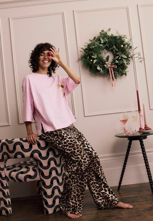 Junge Frau in einem pinken Shirt und einer Hose mit Leopardenmuster sitzt barfuß auf einem gemusterten Stuhl, hält eine Erdbeere vor ihr Auge, neben sich einen Weihnachtskranz und Getränke.