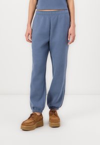 Blaue Sweatpants aus weichem Material, mit elastischem Bund, gerippten Knöcheln und seitlichen Streifen. Kombiniert mit braunen Plattformschuhen.