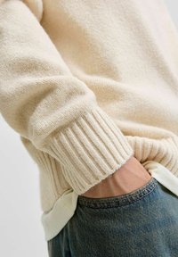 Lys beige strikket sweater med ribbe-ender, parret med blå denim. Blød tekstur tætteste på, der viser sømdetaljer ved kanten og ærmet.