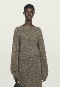 Donna che indossa un maglione oversize marrone e grigio a trama grossa con una gonna coordinata, mentre tiene in mano una piccola borsa scura su uno sfondo semplice.
