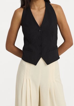 Kobieta w dopasowanej czarnej kamizelce na guziki z klapami oraz kremowych, plisowanych spodniach z wysokim stanem na jednolitym tle.