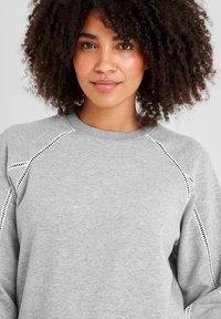 Woman with curly hair wearing a light gray sweatshirt with black and white stitched detailing on the sleeves and shoulders.