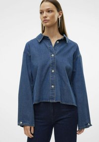 Denim srajca v srednje modri barvi, z ovratnikom, gumbi na sprednji strani in dolgimi rokavi z širokimi manšetami. Ima vidne šive in skrajšan rob.