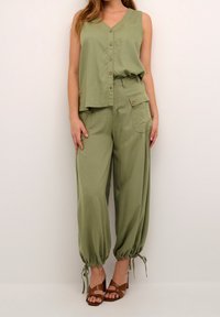 Olivově zelený bezrukávový top s výstřihem do V a předním zapínáním na knoflíky, spárovaný s volnými kalhotami s manžetami, které mají boční kapsy a detaily na zavazování.