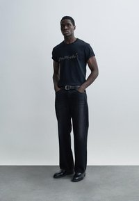T-shirt en coton noir à manches courtes avec un imprimé graphique, associé à un pantalon en denim foncé et des chaussures en cuir noir, debout sur un sol gris.