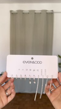 Sada stříbrných náušnic vystavených na růžové kartě, obsahující náušnice s puky, kroužky a visící designy, zdobené různými jasnými rhinestones.