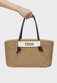 Bolsa de mano de paja trenzada con asas de cuero negro, banda blanca con el logo "TOUS" y detalles de acento anudados. Forma rectangular.
