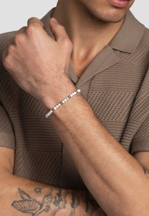 Hvid perlesnor med sølv accenter på et håndled, sat mod en struktureret brun strikket polo shirt. Glatte runde perler og et minimalistisk design.