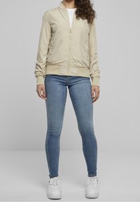 Beige satin bomberjacka med ribbade ärmar och midja, dragkedja fram, och sidofickor, parat med blåa tajta jeans och vita sneakers.