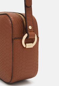 Borsa in pelle marrone con texture, dotata di una cerniera dorata, anello decorativo e etichetta del marchio. Presenta una forma strutturata e un dettaglio di cucitura a contrasto.
