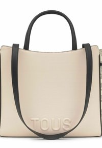 Bolso tote beige con asas negras y correa para el hombro, logo "TOUS" en relieve en el frente, y un panel lateral con patrón visible en el lado derecho.
