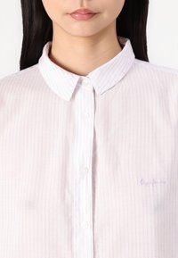 Camisa abotoada de riscas roxas claras com colarinho espalhado, apresentando botões brancos e um discreto logo bordado no peito.