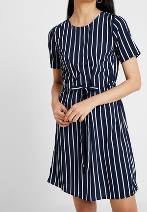 Vrouw draagt een marineblauwe jurk met witte verticale strepen, korte mouwen en een gestrikte ceintuur in de taille.