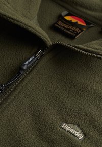 Groene fleecejas met een voorrits. Heeft een gestructureerd oppervlak, een logopatch en een zwart koordje aan de rits.