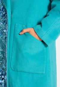 Türkiser Wollmantel mit einer großen Seitentasche, einer weichen, strukturierten Oberfläche und einem glänzenden, mit Pailletten ausgekleideten Innenfutter.