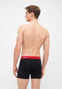 Čierne boxerky s červeným elastickým pásom, na ktorom je logo "HUGO", zobrazené zozadu bez ďalšieho oblečenia.