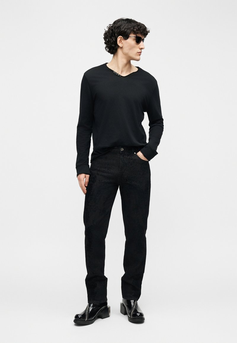 Uomo con indosso una maglietta nera a maniche lunghe, pantaloni neri con trama, stivali neri lucidi e occhiali da sole scuri, che guarda di lato con una mano in tasca.