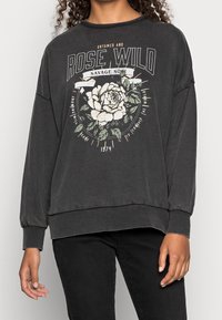 Sudadera gris oscuro con un gráfico floral en blanco y verde, y texto "ROSE WILD". Corte holgado y mangas largas. Textura suave.