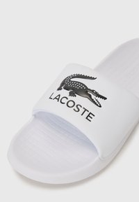 Sandale blanche à glissement avec une bande structurée, arborant un logo crocodile noir et le texte "LACOSTE". Semelle texturée pour une meilleure adhérence.