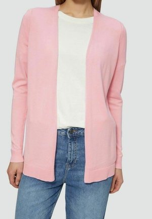 Vrouw draagt een lichtroze open voor vest over een wit shirt en blauwe spijkerbroek, staand tegen een effen achtergrond.
