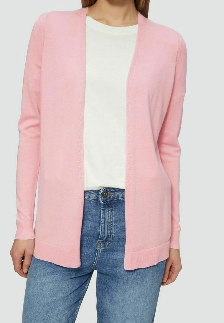 Femme portant un cardigan rose pâle ouvert sur un t-shirt blanc et un jean bleu, debout devant un fond uni.