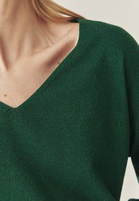 Zaļš V-veida kakla džemperis no spīdīga trikotāžas auduma. Raksturots ar gludu tekstūru, pieguļošu dizainu un plašu kakla izgriezumu.