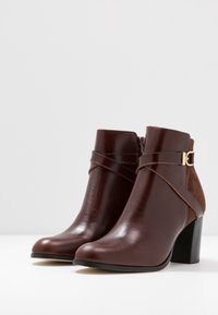Bottes courtes en cuir marron à finition brillante, accents en daim et boucle dorée. Bout carré et talon empilé.