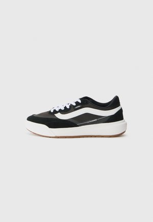 Black and gray sneakers with white accents, featuring a textured suede upper, white laces, and a rubber gum sole. Notable brand logo visible.