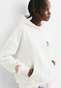Witte hoodie met een vo zak en ribgebreide manchetten. Heeft zwarte tekst op de borst en rode tekst op de rechtermouw.