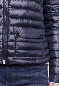 Lähedalt näha mereväe sinine tikitud jope, millel on tõmblukk ja nööbiga tasku, kandmiseks siniste teksade ja heleda särgi manseti kohal.