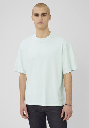 Jeune homme aux cheveux blonds courts portant un t-shirt oversize bleu clair et un jean foncé sur un fond gris uni.