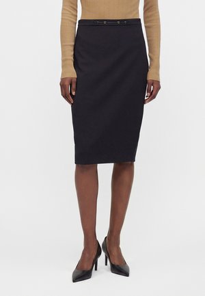 Femme portant une jupe crayon noire longueur genoux, un top à manches longues couleur beige et des chaussures à talons noirs, se tenant devant un fond uni.