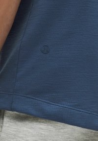 Detailný záber na lem modrého športového trička s malým okrúhlym logom Lululemon nad šedým látkovým opaskom.