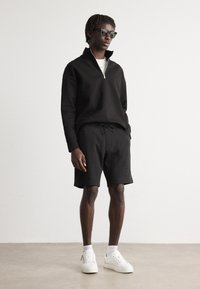 Felpa nera con zip e collo alto, pantaloni corti neri, sneaker bianche indossate con calze. Presenta una vestibilità rilassata e un design minimalista.