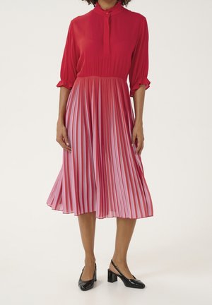 Kobieta ubrana w czerwoną bluzkę z rękawami 3/4 i plisowaną różową spódnicę z wysokim stanem, do tego czarne sandały na słupku z paskiem z tyłu.