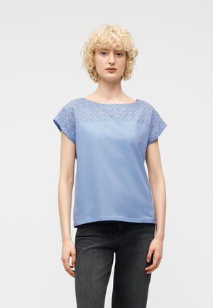 Žena s kučeravými blond vlasmi, oblečená v svetlomodrom tričku s krátkym rukávom a ozdobou z dierkovaného vzoru, a čiernych džínsach, stojaca pred jednofarebným pozadím.