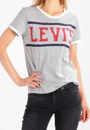 Osoba ubrana w szarą koszulkę Levi's z czerwonym logo i granatowymi paskami, w połączeniu z czarnymi dżinsami, pozująca z jedną ręką na biodrze.