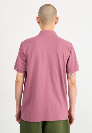 Person med barberet hoved iført en støvet lyserød kortærmet polo og grønne bukser, set bagfra, med en tatovering på armen.