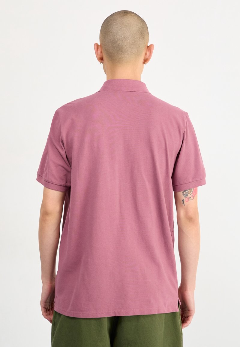 Personne au crâne rasé portant un polo à manches courtes rose poussiére et un pantalon vert, vue de dos, montrant un tatouage sur le bras.