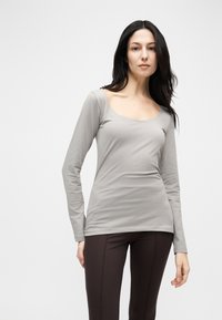 Γυναίκα με μακριά σκούρα μαλλιά, που φοράει ανοιχτό γκρι μπλούζα με μακριά μανίκια και στρογγυλή λαιμόκοψη, καθώς και σκούρο εφαρμοστό παντελόνι, στέκεται μπροστά σε λευκό απλό φόντο.