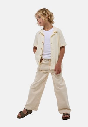 Chemise beige clair à manches courtes et boutonnée sur un t-shirt blanc, associée à un pantalon large beige et des sandales brunes. Tissu à texture lisse.