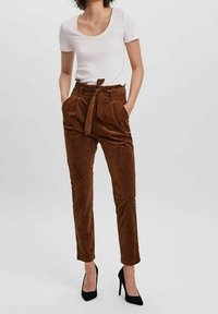 Kvinna klädd i bruna högmidjade byxor i kordflorsmaterial med knytbälte, en vit kortärmad topp och svarta högklackade skor, ståendes med händerna i fickorna.