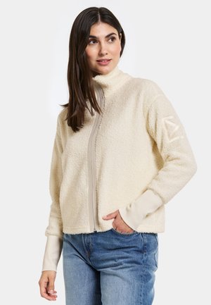 Veste en polaire couleur crème avec un col montant, fermeture éclair intégrale et texture douce. Présente un devant beige, des poignets extensibles et un détail de logo sur la manche.