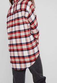 Camicia di flanella a quadri rossi, blu e crema con orlo arrotondato. Maniche lunghe e dettaglio alla yoke sulla parte posteriore, abbinata a jeans scuri.
