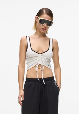 Naine kannab ülemõõdulisi valgeid päikeseprille, valget kurrutatud lühikeset pluusi musta äärega ja musti paelaga pükse, mille taskul on logotekst.