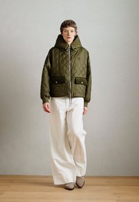 Khakifarbene gesteppte Jacke mit großer Kapuze, Frontreißverschluss und zwei Fronttaschen; kombiniert mit weißen weit geschnittenen Hosen und grauen Stiefeletten.