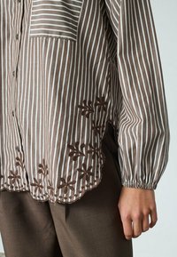 Brun og hvidstribet skjorte med knapper og en bølget kant, der har blomsterbroderi og en lomme på venstre side. Lange, rynkede ærmer.