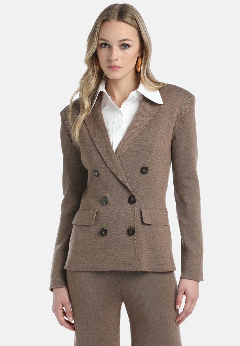 Vrouw in een beige dubbelzijdig colbert en bijpassende broek over een wit overhemd, staand tegen een effen lichte achtergrond.