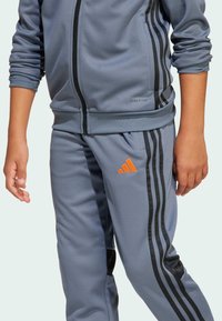 Niño vestido con un chándal azul grisáceo con rayas negras, logotipo naranja en los pantalones, mangas remangadas y manos relajadas a los lados.